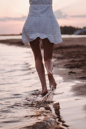 Close-up of a running woman. Photo of legs back view of a beautiful tanned woman in a white summer dress on the river bank against the background of sunset and cloudsの写真素材
