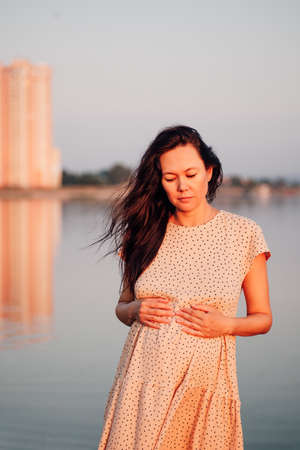Brunette on background of urban landscape. Beautiful long-haired Asian woman in silk polka dot dress is thinking and admiring sunset on seashoreの写真素材