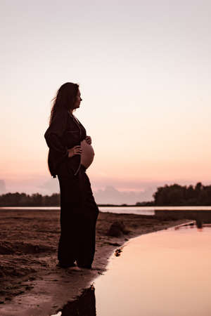 Pregnant woman on beach at sunset. Silhouette in profile against background of setting sun of beautiful young brunette with long hair and dark summer suit enjoying warm summer evening in natureの写真素材