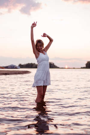 A young woman is dancing in the water. Photo a beautiful happy blonde in a white summer dress and with her hands raised up dancing against the sunset backgroundの写真素材