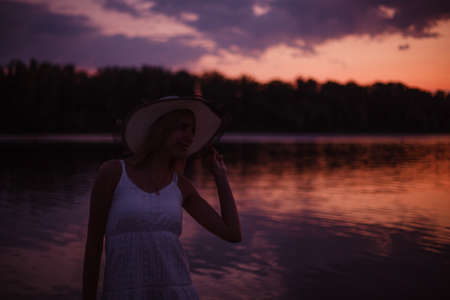 A woman in a straw hat is standing by the river. A beautiful young slender blonde in a white lace dress poses against the sunset and holds a hat her handの写真素材