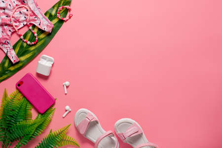 Summer beach top view concept. Children summer clothing and accessories on the pink background with green leavesの写真素材