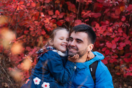 close up happy family portrait. happy smiling father man with dark hair hold his smiling little daughter in blue jacket at autumn forest backgroundの写真素材