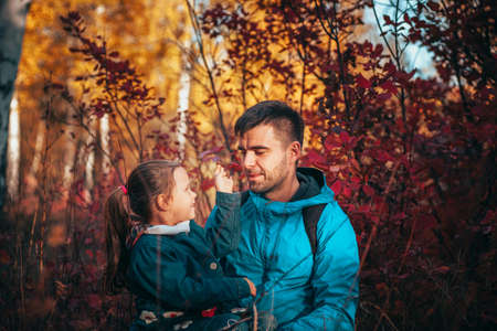 close up happy family portrait. man with dark hair look at his little daughter in blue jacket at autumn forest backgroundの写真素材