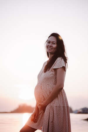 Pregnant woman smiles. Beautiful young woman in summer dress and long brown hair stands on beach, looks at camera and holds her stomach with her handsの写真素材