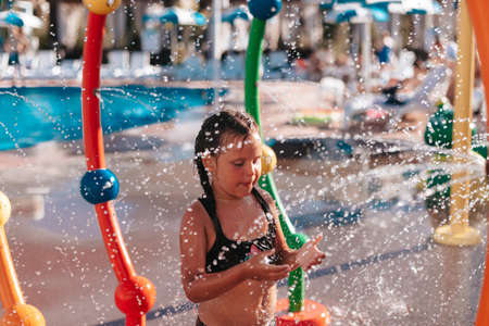 Child is playing by pool. Little girl in black swimsuit is sitting by pool and playing with water...