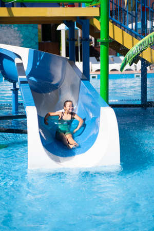 Cute girl is riding water slide. Child spends sunny summer day in water park at tropical resort by...
