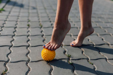 details of young woman bare feet standing on tiptoes on spiky rubber yellow ball to relax muscles and reduce pain on paving tile on sunny dayの写真素材