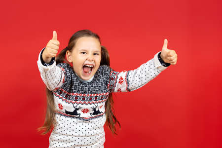 portrait of laughing, joyful girl in white Christmas sweater with reindeer giving thumbs up,...
