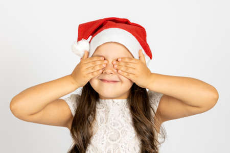close-up portrait of smiling girl with long curls in white dress, in red Santa hat, covering eyes with palms, isolated on white background, concept of waiting for surpriseの写真素材