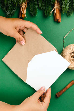 top view of Christmas card on craft envelope, woman hands, green table and Christmas decorationsの写真素材