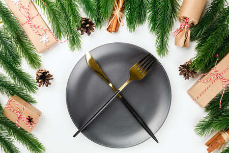 golden knife and fork crossed on black plate on Christmas dinner table with frame of fir branches, with cones and giftsの写真素材