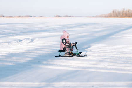 Child rides snowmobile. Little girl in pink warm jacket enjoys walk in nature and sledding on frozen river on sunny winter dayの写真素材