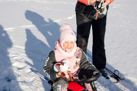 Little girl enjoys walk. Child squints from sun and enjoys sledge ride through snowy forest, shadows on snowの写真素材