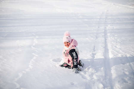 Little girl is sitting in snow. Child in pink winter jacket and hat with pompom has fun on walk in nature and enjoys sunny weather and plays with snow on frosty winter morningの写真素材