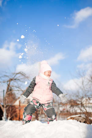 Girl is playing snowballs against blue sky. Child in warm winter suit has fun, plays and jumps on snowdrifts, rides snow slide while walking in natureの写真素材
