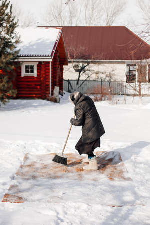 Side view of woman cleaning carpet. Caucasian woman is engaged in traditional method of ecological carpet cleaning with fresh snow and broom in backyard on background of wooden cottageの写真素材