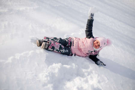 Child is playing with snow. Little girl enjoys walk in nature, having fun and riding snow slide without sled on sunny winter dayの写真素材