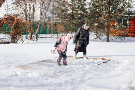 Woman and child are cleaning carpet. Family is engaged in traditional method of ecological carpet cleaning with help of fresh snow and carpet beater in backyard near wooden cottageの写真素材