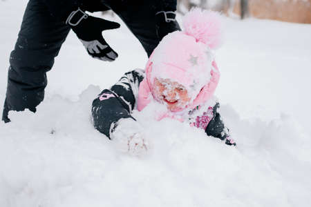 kid lying in snowdrift wearing pink winter clothes and father helping daughter to stand up in...
