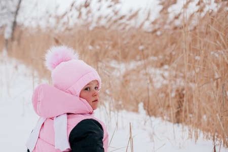Closeup photo of female Russian kid with back to camera sitting in snow and looking behind wearing pink winter clothes in forest. Astonishing background full of white color and snowの写真素材