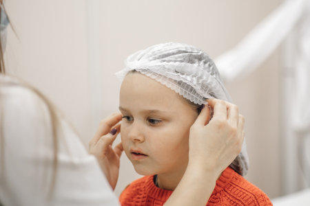 close-up of beautician hands putting white disposable medical cap on head of six-year-old girl in...