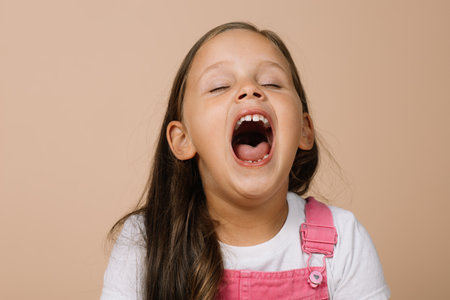 Little girl with fully opened mouth and closed eyes yawning looking tired wearing bright pink...