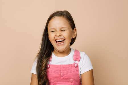 Portrait of child with fully opened mouth, closed eyes and excited smile with teeth looking at camera wearing bright pink jumpsuit and white t-shirt on beige backgroundの写真素材