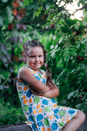 Nice little girl sitting on log looking at camera smiling with folded arms and pigtail on head wearing light summer dress. Charming background full of greenの写真素材