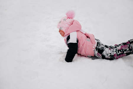 Closeup photo of female kid lying and crawling in snow looking away wearing pink winter clothes in forest. Astonishing background full of white color and snowの写真素材