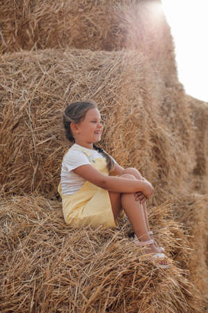 Portrait of sitting girl on high haystack smiling with teeth looking somewhere far away wearing sundress. Having fun away from city on field full of golden hayの写真素材