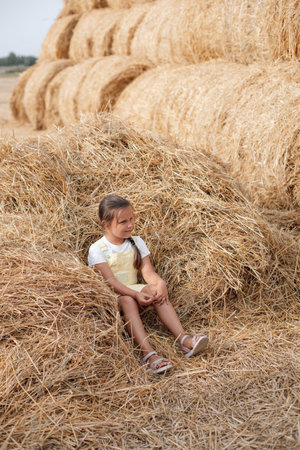 Big pile of hay harvest on field with young girl resting in it looking away with serious face holding hay in hand wearing sundress. Having fun away from city on field full of golden hayの写真素材
