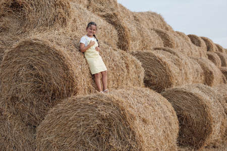 Smiling young girl in sundress standing on haystack leaning on another haystack looking at camera...