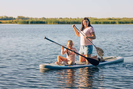 Happy Caucasian mother and little daughter sup boarding with oars in hands looking at camera and...