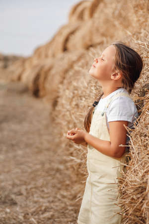 Serious little girl leaning on high haystack in full of hay field with braid on shoulder holding hands near belly with head up and closed eyes. Time away from city in country fieldの写真素材