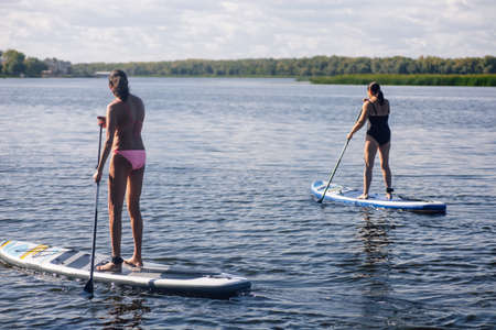 Two middle aged women puddle boarding on beautiful blue lake with oars in hands with trees and reeds...