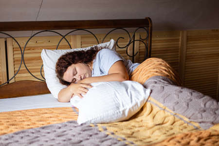 Upset curly brunette female adult in bed under terry covers with closed eyes hugging pillow wearing...