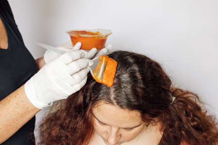 Lady with dyeing brush in one hand and bowl in the other putting red substance on woman hair on white background. Hair coloring at home. Getting rid of gray hair. Anti-ageの写真素材