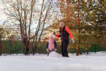 Cute small daughter and mother rolling big snow balls for constructing snowman smiling on backyard in evening with rowan and fir trees in background. Parents spending time with childrenの写真素材