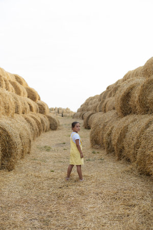 Serious young Caucasian girl standing in field with feet covered with hay with back to camera and...