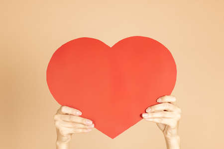 Female hands holding heart shaped red paper postcard on background of skin color. Celebrating and...