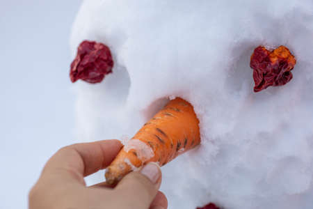 Close up of hand making snowman putting carrot as nose on face with red eyes on backyard in daytime while having walk. Parents spending time with childrenの写真素材
