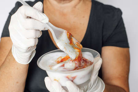 Old woman holding small transparent bowl with hair dye substance and long plastic brush for hair coloring in hands covered with white latex gloves. Anti ageの写真素材