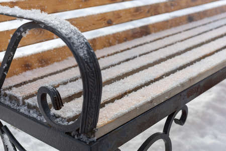 Close up of wooden bench with black colored iron frame slightly covered with snow put somewhere in...