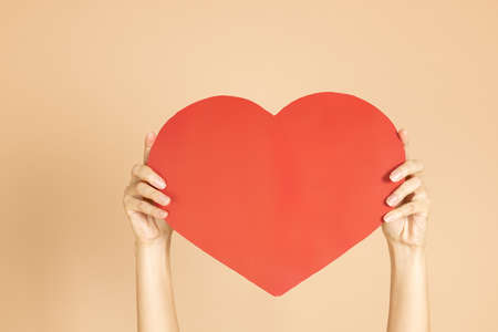 Closeup of woman hands having beautiful red paper heart on body colored background. Celebrating and congratulating loved one with Valentines day. Expressing loveの写真素材