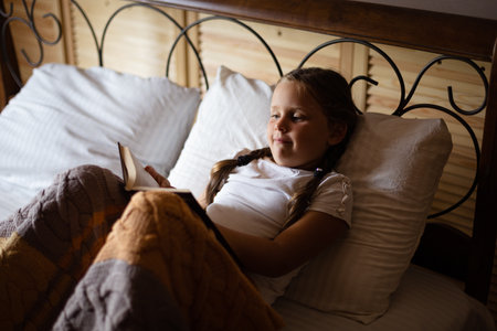 Curious little female child lying on huge double bed with book in hands looking at book and reading wearing pajamas in wooden country house. Leisure time activitiesの写真素材