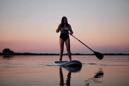 Female sup boarder holding oar in hands rowing looking at camera on calm lake with stunning pink sky in background in swimsuit in summer. Active lifestyle for older peopleの写真素材
