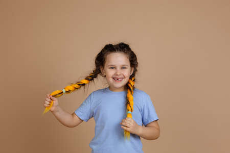 Funny cheerful joyful female child with missing tooth smiling looking at camera holding yellow kanekalon braids with hands on beige background wearing blue t-shirt and jeansの写真素材