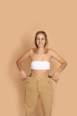 Portrait of smiling woman in good shape in oversize pants, white underwear showing slim waist by pulling pants on skin colored background. Weight loss. Keeping a healthy diet, exercisingの写真素材