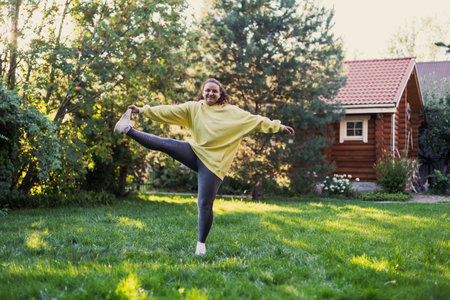 Female adult smiling on green grass doing exercises in sportswear with one foot up on backyard full of sun light with country house and high trees in background. Body positiveの写真素材